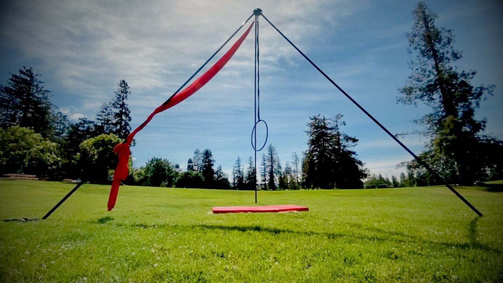 Freestanding aerial rig set up on a park lawn with red silks and a lyra