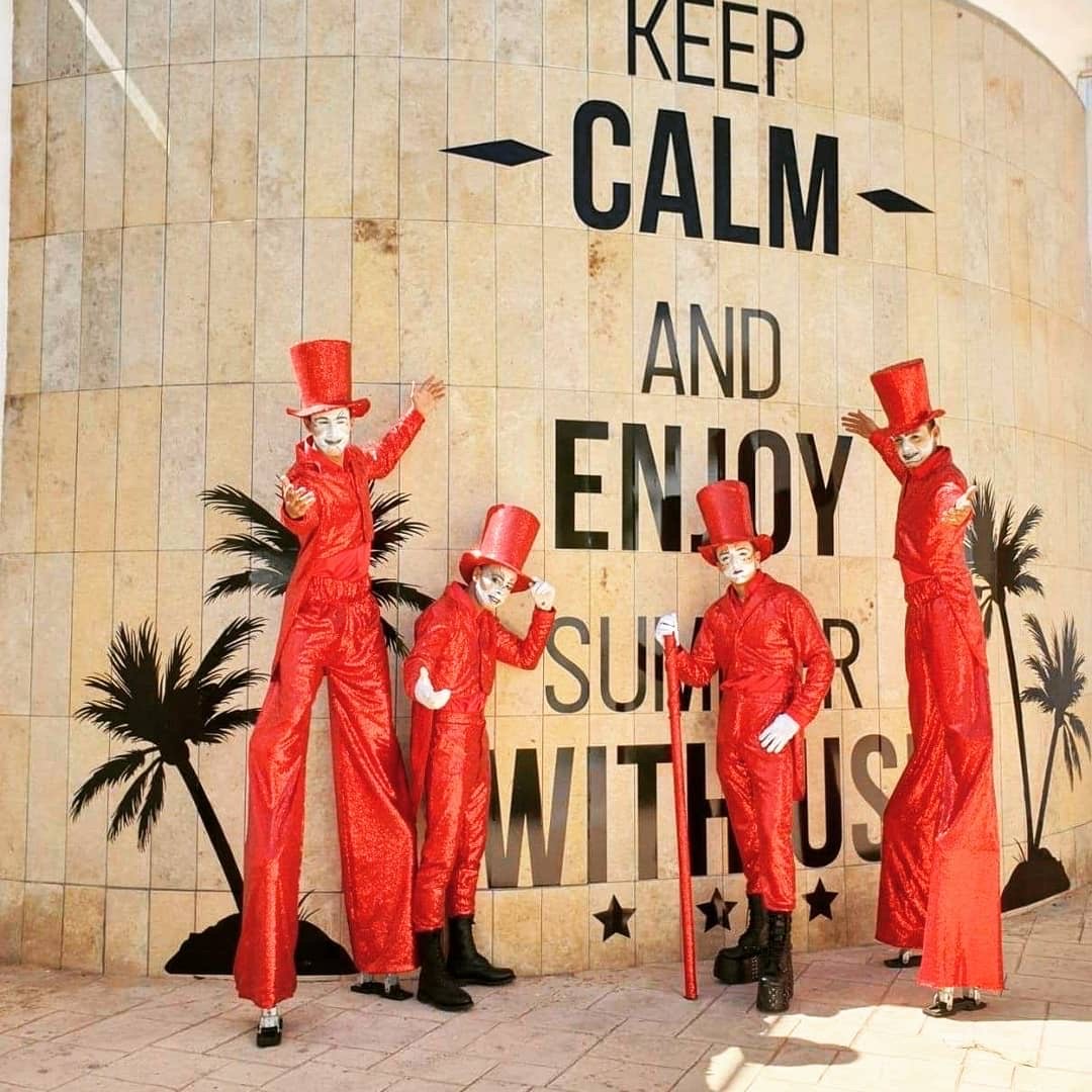 Four stilt walkers in sparkly red suits and top hats posing in front of a wall